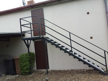 Zewnętrzne schody metalowe z platformą prowadzące do drzwi na piętrze budynku, widoczne z boku na tle jasnej elewacji i żwirowego podłoża.