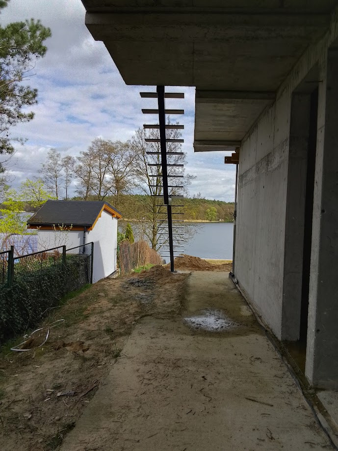 Surowe schody metalowe przymocowane do betonowej ściany budynku w stanie surowym, z widokiem na jezioro i zieleń w tle.