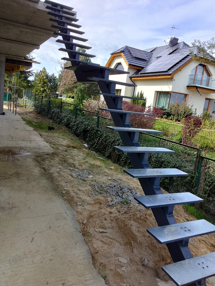 Surowe, zewnętrzne schody metalowe z płaskimi stopniami, prowadzące do niezadaszonego piętra budynku w trakcie budowy, z domem jednorodzinnym w tle.