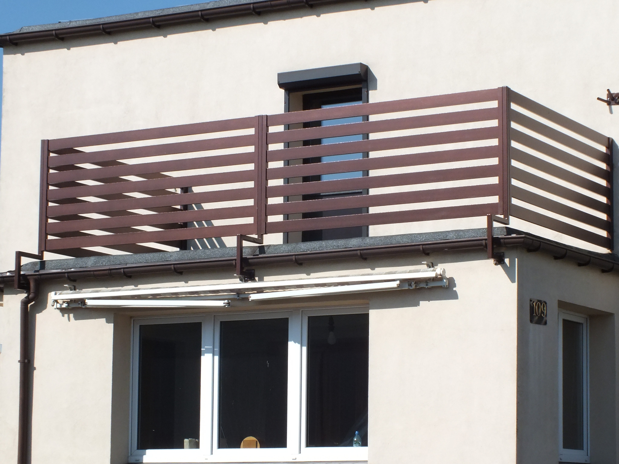 Balkon z nowoczesną, poziomą balustradą w kolorze brązowym, widok z dołu na elewację budynku z oknami i roletami.