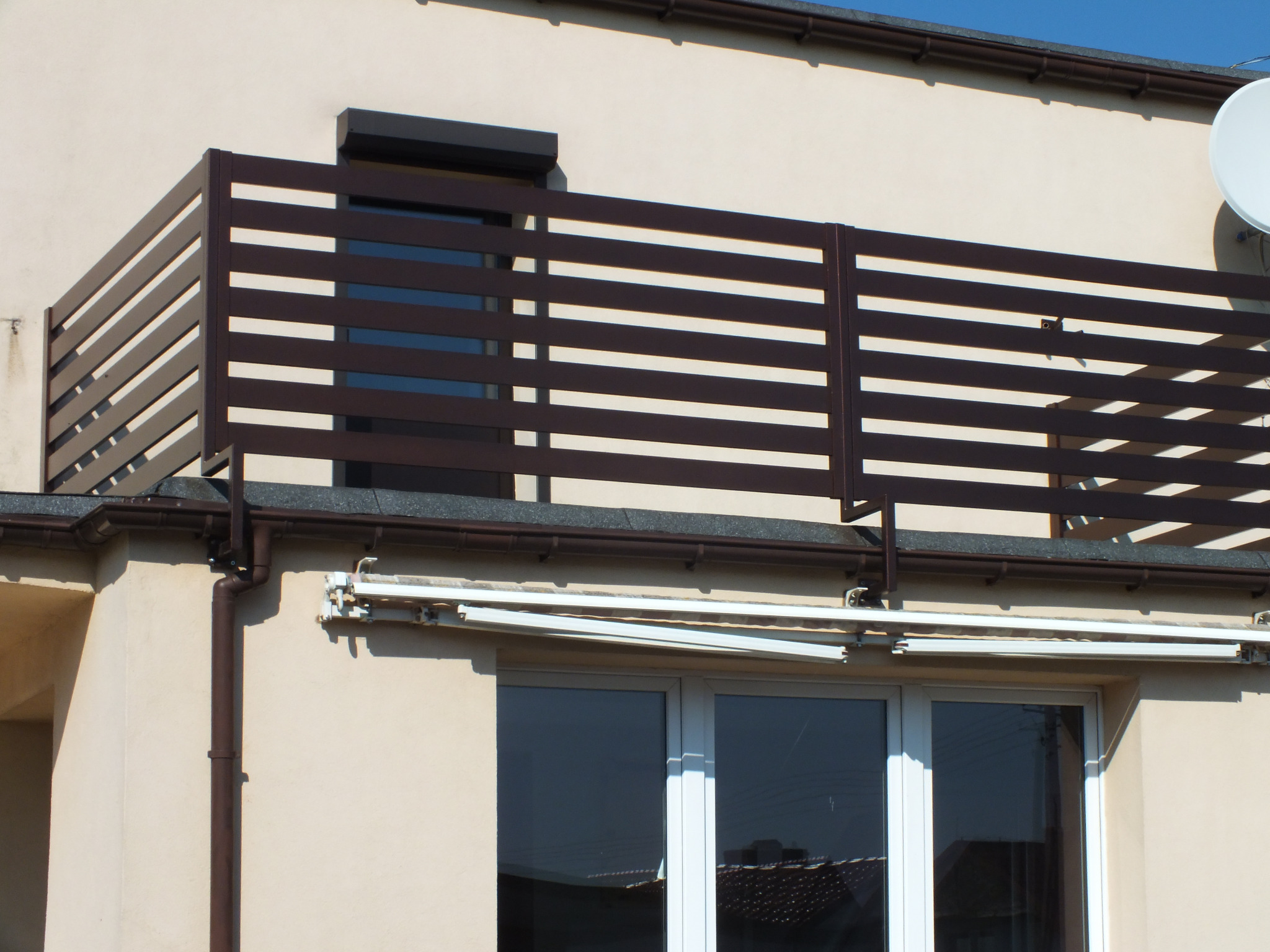 Brązowa balustrada balkonowa z poziomych listew na tle jasnobeżowej elewacji budynku z widocznym oknem i anteną satelitarną.