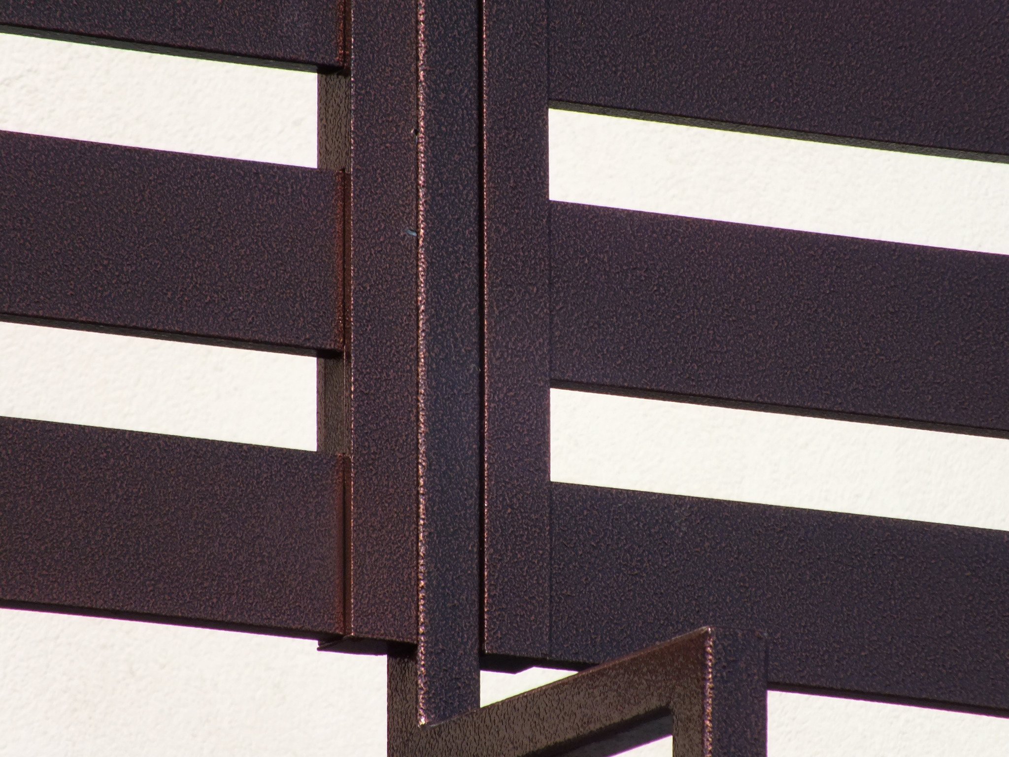 Fragment metalowej konstrukcji z poziomymi, brązowymi profilami na tle białej ściany, widoczne detale tekstury powierzchni.