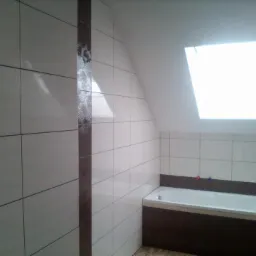 Biała łazienka na poddaszu z oknem dachowym, wykończona jasnymi płytkami z brązowym pasem dekoracyjnym, obudowana wanna.