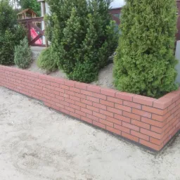 Obrzeże rabaty kwiatowej z czerwonej cegły klinkierowej, otaczające zielone tuje, na piaszczystym podłożu, z widocznym ogrodzeniem w tle.