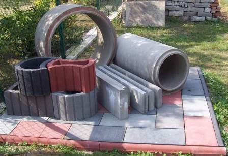 Ekspozycja różnorodnych elementów betonowych: kręgi, rury, obrzeża, krawężniki, gazony i płytki chodnikowe ułożone na palecie z kostki brukowej, na tle zieleni.