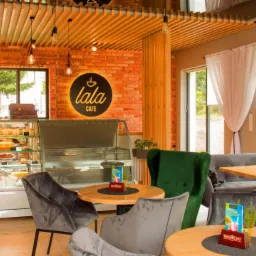 Wnętrze kawiarni w stylu loft z ceglaną ścianą, drewnianym sufitem, witryną z ciastami i stolikami z aksamitnymi krzesłami.