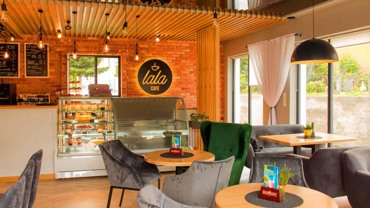 Wnętrze kawiarni w stylu loft z ceglaną ścianą, drewnianym sufitem, witryną z ciastami i stolikami z aksamitnymi krzesłami.