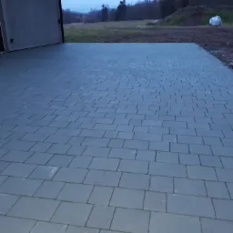 Nowo ułożona, szara kostka brukowa przed budynkiem z zamkniętymi roletami, widoczne palety z boku i worki w tle.