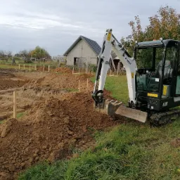 Mini koparka Bobcat E19 na trawiastym terenie, obok wykopu pod fundamenty z drewnianymi szalunkami, w tle budynek gospodarczy i drzewa.