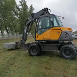 Żółto-szara koparka Mecalac 714MW na trawiastym terenie, podnosi paletę z betonowymi elementami, w tle ogrodzenie i drzewa.