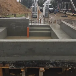 Surowy, betonowy fundament z wypełnieniem wodą, widoczne zbrojenie i deskowanie, w tle plac budowy z hałdą ziemi i budynkiem w stanie surowym otwartym.