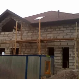 Surowy budynek w trakcie wznoszenia z bloczków, z widoczną konstrukcją dachu pokrytą brązowym gontem bitumicznym i oknem połaciowym; na pierwszym planie kontener budowlany i mieszalnik do betonu.