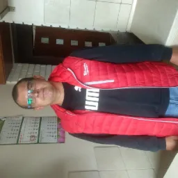 Mężczyzna w czerwonej kamizelce Milwaukee, czarnej koszulce Puma i okularach, na tle kalendarza z datą 16 września 2019, w kuchni z brązowymi szafkami.