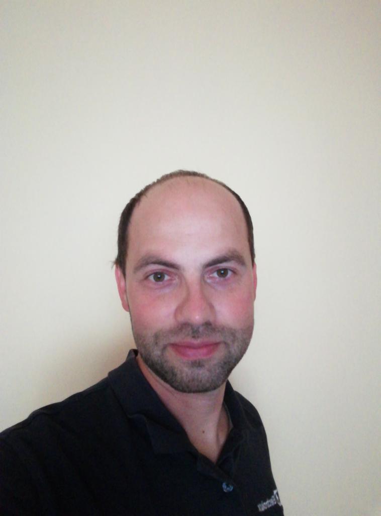 Portret mężczyzny z krótką brodą i ciemnych włosach na jasnym tle, ubrany w ciemną koszulkę polo z widocznym logo firmy.