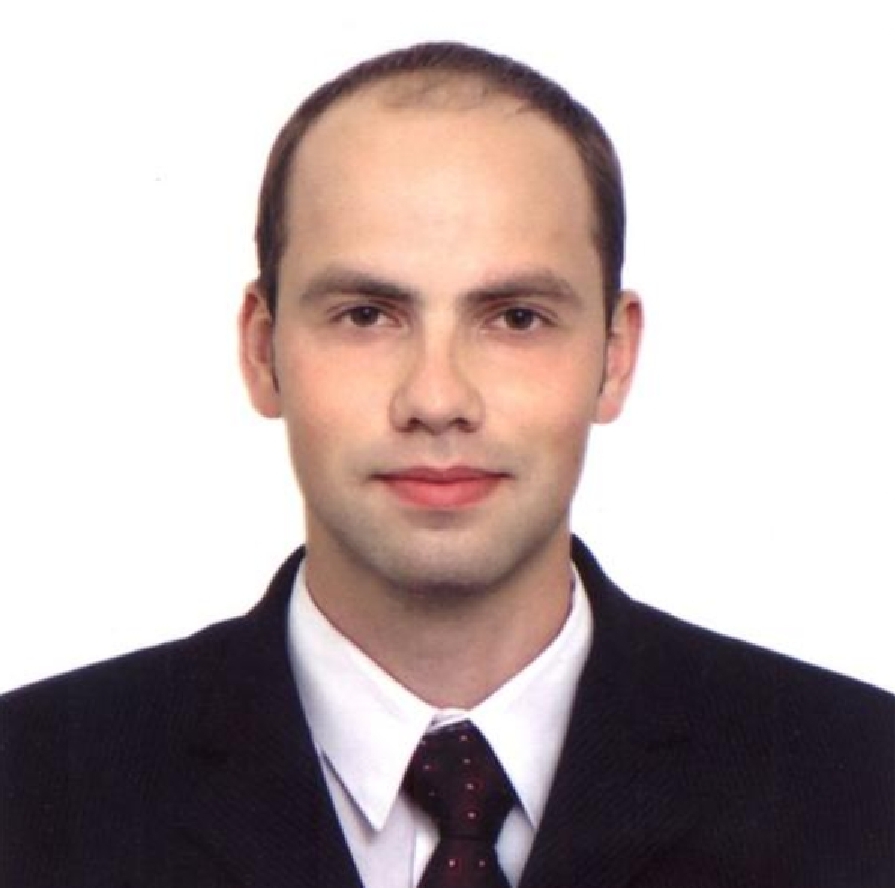 Portret mężczyzny w garniturze na białym tle. Ujęcie od przodu, widoczna twarz i część torsu. Ciemny garnitur, biała koszula, bordowy krawat w kropki.