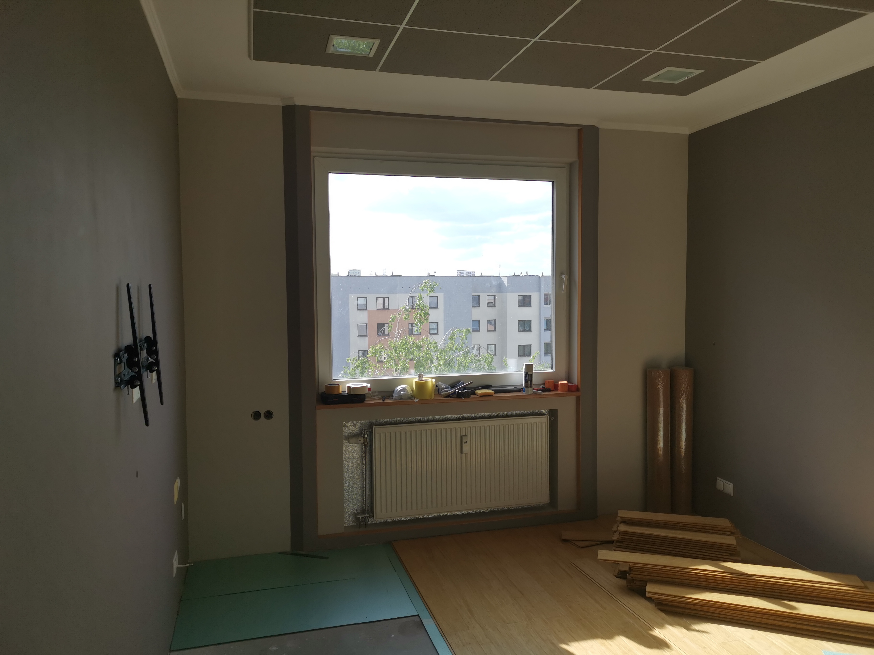 Wnętrze w trakcie remontu: panele podłogowe ułożone częściowo, widoczny grzejnik, okno z widokiem na blok, narzędzia na parapecie, przygotowane rolki podkładu pod panele, ściany w dwóch odcieniach...