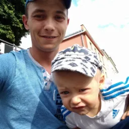 Selfie mężczyzny z synem w czapce z motywem Batmana na tle budynku z czerwonym dachem i fragmentem zieleni.