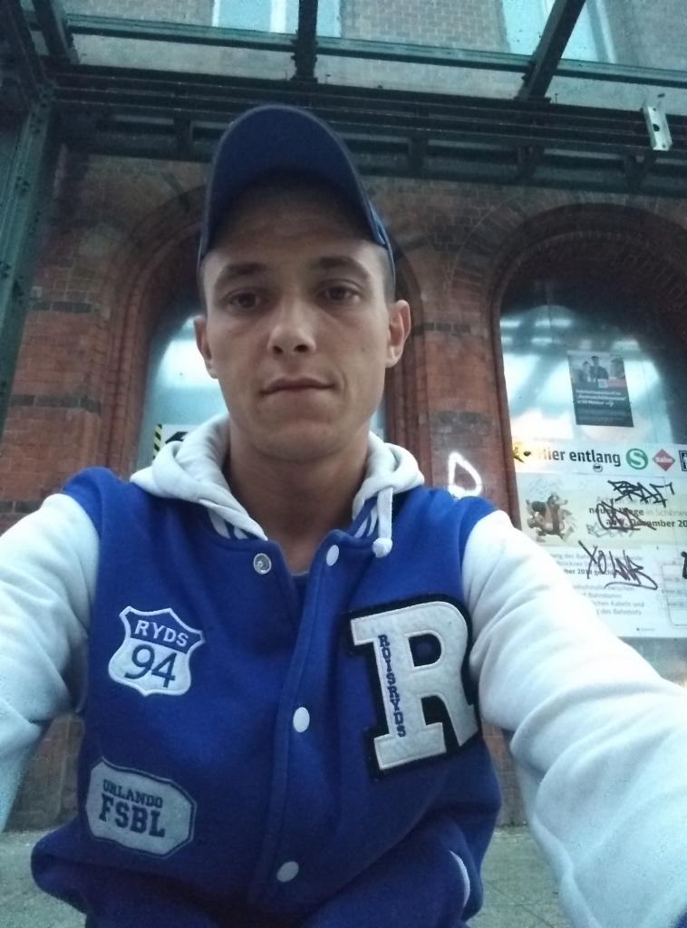 Mężczyzna w bluzie bejsbolówce i czapce z daszkiem na tle ceglanej ściany z łukowatymi oknami i informacjami. Ujęcie z dołu, oświetlenie mieszane.