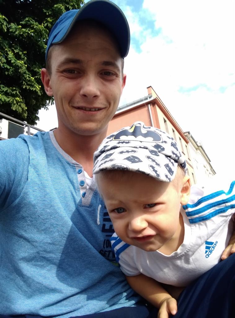 Selfie mężczyzny z synem w czapce z motywem Batmana na tle budynku z czerwonym dachem i fragmentem zieleni.