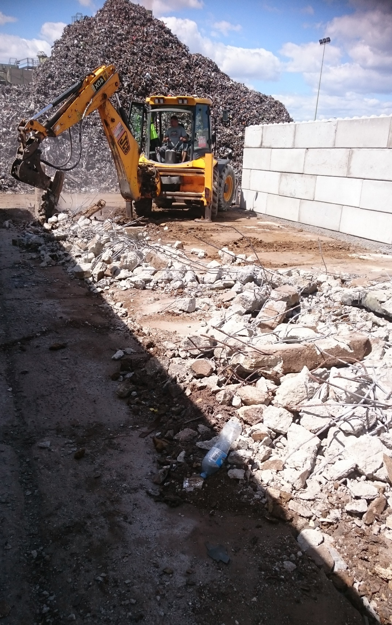 Żółta koparka JCB kruszy betonowe elementy na placu budowy z widoczną górą gruzu i plastikową butelką w pierwszym planie.
