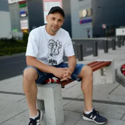 Mężczyzna w czapce siedzi na ławce przed budynkiem z logotypami, ubrany w białą koszulkę i dżinsowe spodenki, z rękami splecionymi na kolanach.