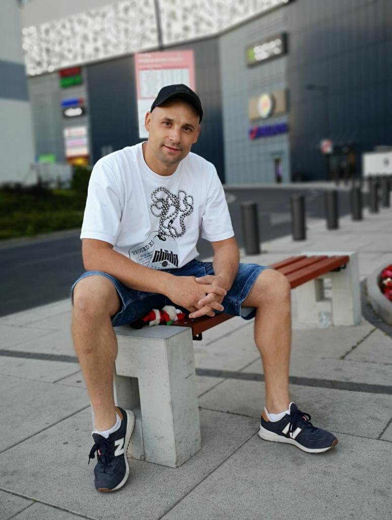 Mężczyzna w czapce siedzi na ławce przed budynkiem z logotypami, ubrany w białą koszulkę i dżinsowe spodenki, z rękami splecionymi na kolanach.