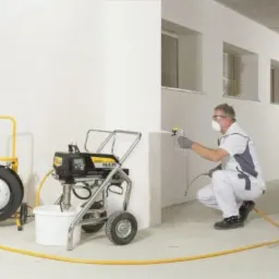 Pracownik w kombinezonie i masce maluje ścianę w pomieszczeniu za pomocą agregatu malarskiego Wagner PS3-29, widoczne okna w stanie surowym.