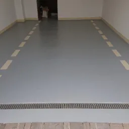 Szara, gładka posadzka z liniami w kolorze ecru, wyznaczającymi krawędzie, oraz metalowa kratka odpływowa przy wejściu do pomieszczenia.