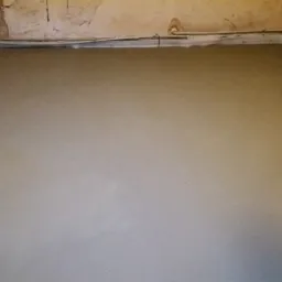 Gładka, szara posadzka jastrychowa w pomieszczeniu z widocznymi rurami przy ścianie i fragmentem czarnej ramy okiennej w prawym rogu.