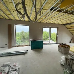 Poddasze w trakcie ocieplania wełną mineralną, widoczne profile metalowe, okna dachowe i worki z gładzią szpachlową.