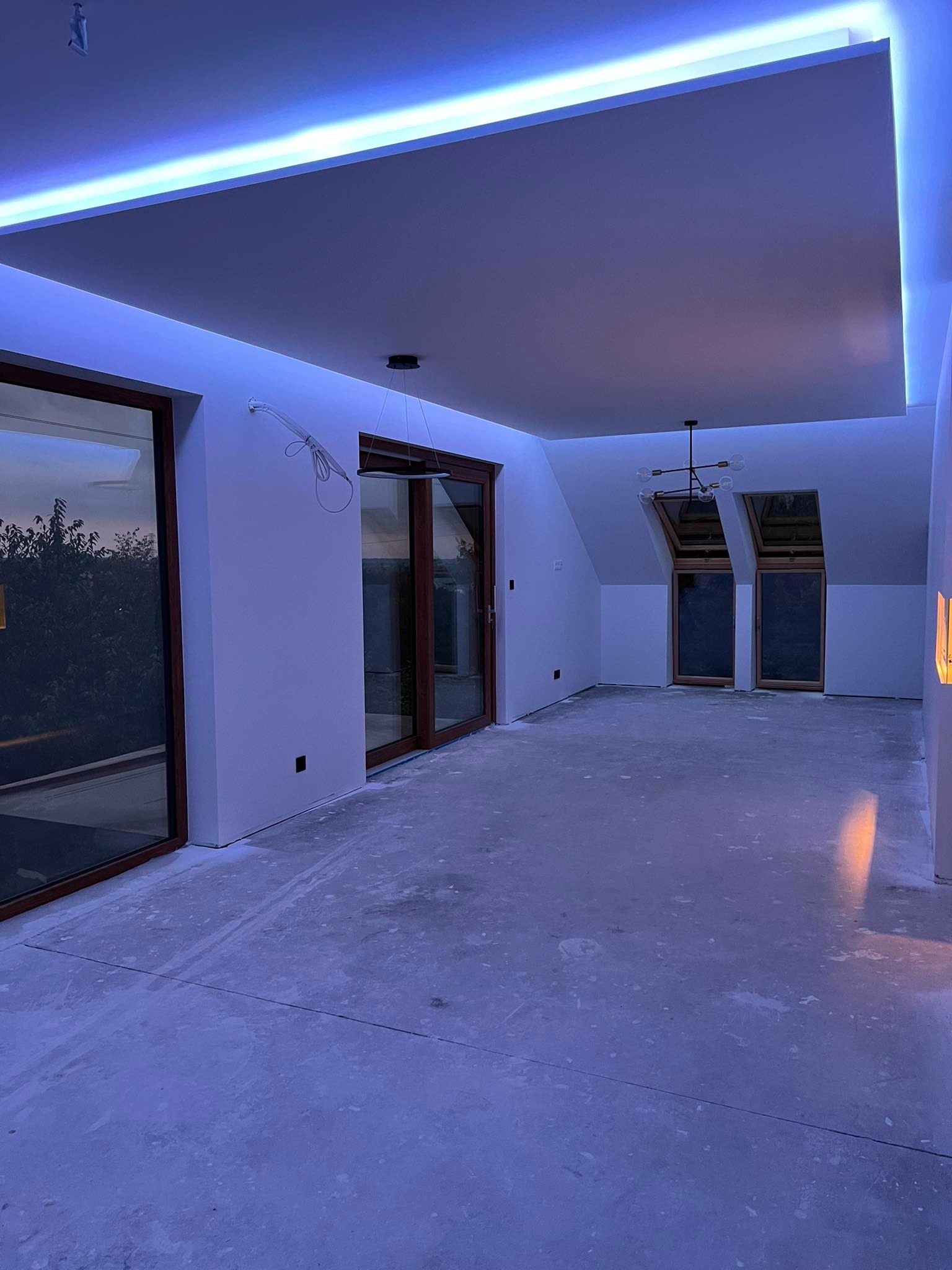 Wnętrze pokoju po remoncie z podwieszonym sufitem z oświetleniem LED, widoczne okna dachowe i duże przesuwne okna balkonowe, ściany pomalowane na biało, na podłodze widoczne ślady po pracach...