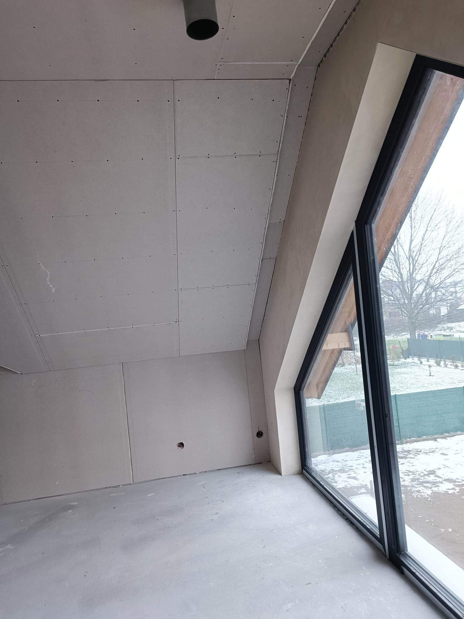 Poddasze w trakcie prac wykończeniowych: zamontowane płyty na skosach i suficie, widoczne otwory pod instalacje, duże okno z widokiem na zaśnieżony ogród.