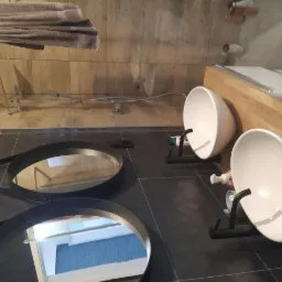 Dwie nowoczesne, białe umywalki nablatowe na czarnych, minimalistycznych stelażach, zamontowane obok blatu z jasnego drewna, z okrągłymi lustrami w czarnych ramach leżącymi na ciemnej, grafitowej...