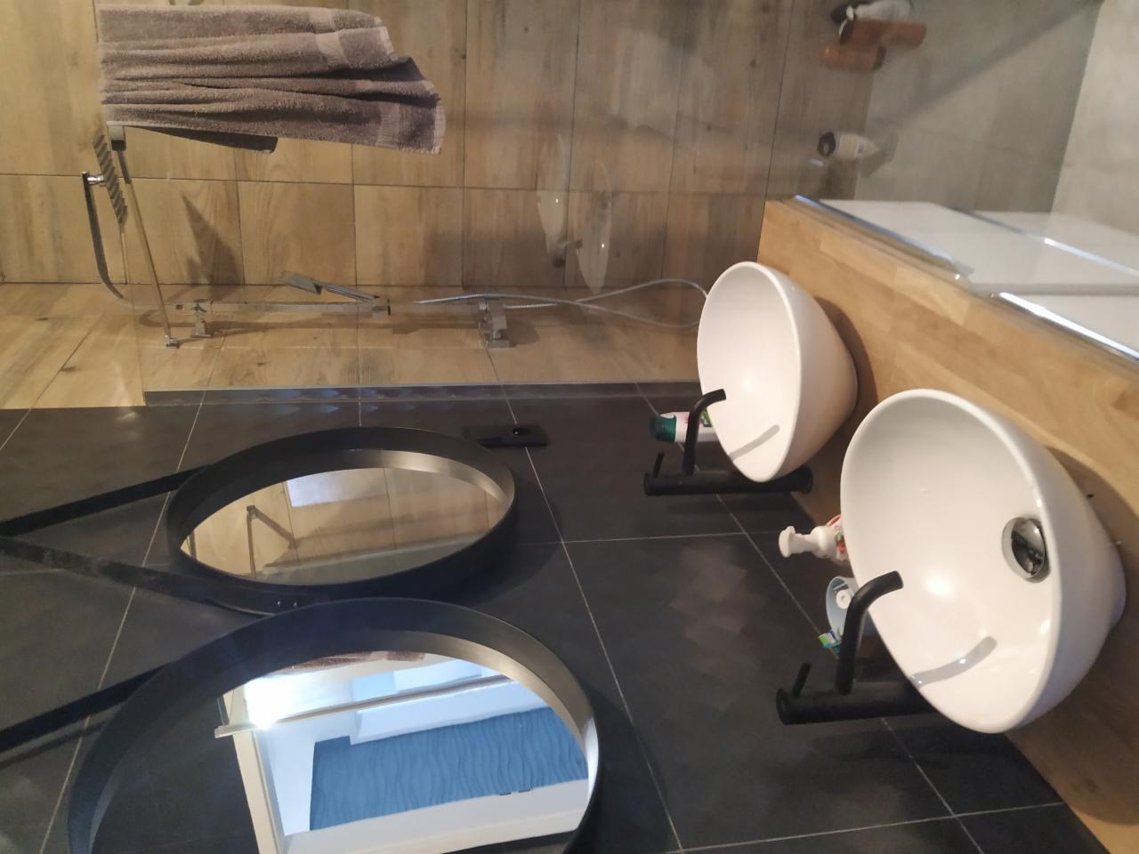 Dwie nowoczesne, białe umywalki nablatowe na czarnych, minimalistycznych stelażach, zamontowane obok blatu z jasnego drewna, z okrągłymi lustrami w czarnych ramach leżącymi na ciemnej, grafitowej...