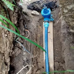 Niebieski zawór i rura wodna zamontowane w wykopie, podparte drewnianą deską, widoczne korzenie i ziemia.