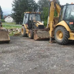 Trzy żółte koparko-ładowarki CAT 428E zaparkowane na żwirowym placu, widok z przodu, w tle dom i góry.