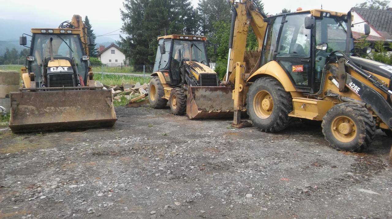 Trzy żółte koparko-ładowarki CAT 428E zaparkowane na żwirowym placu, widok z przodu, w tle dom i góry.