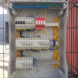 Otwarta skrzynka elektryczna z zamontowanymi bezpiecznikami, ogranicznikami przepięć i listwami zaciskowymi, widoczna na tle metalowego ogrodzenia i budynku.