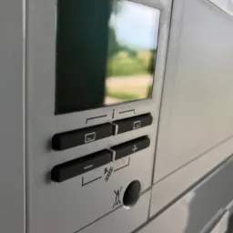 Panel sterowania urządzenia grzewczego z wyświetlaczem i przyciskami regulacji, widoczne odbicie otoczenia w wyświetlaczu.