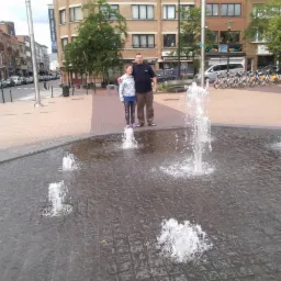 Para stoi na fontannie w mieście. W tle budynki, rowery miejskie i zaparkowane samochody. Ujęcie z poziomu fontanny, woda w ruchu. Lekkie zachmurzenie.