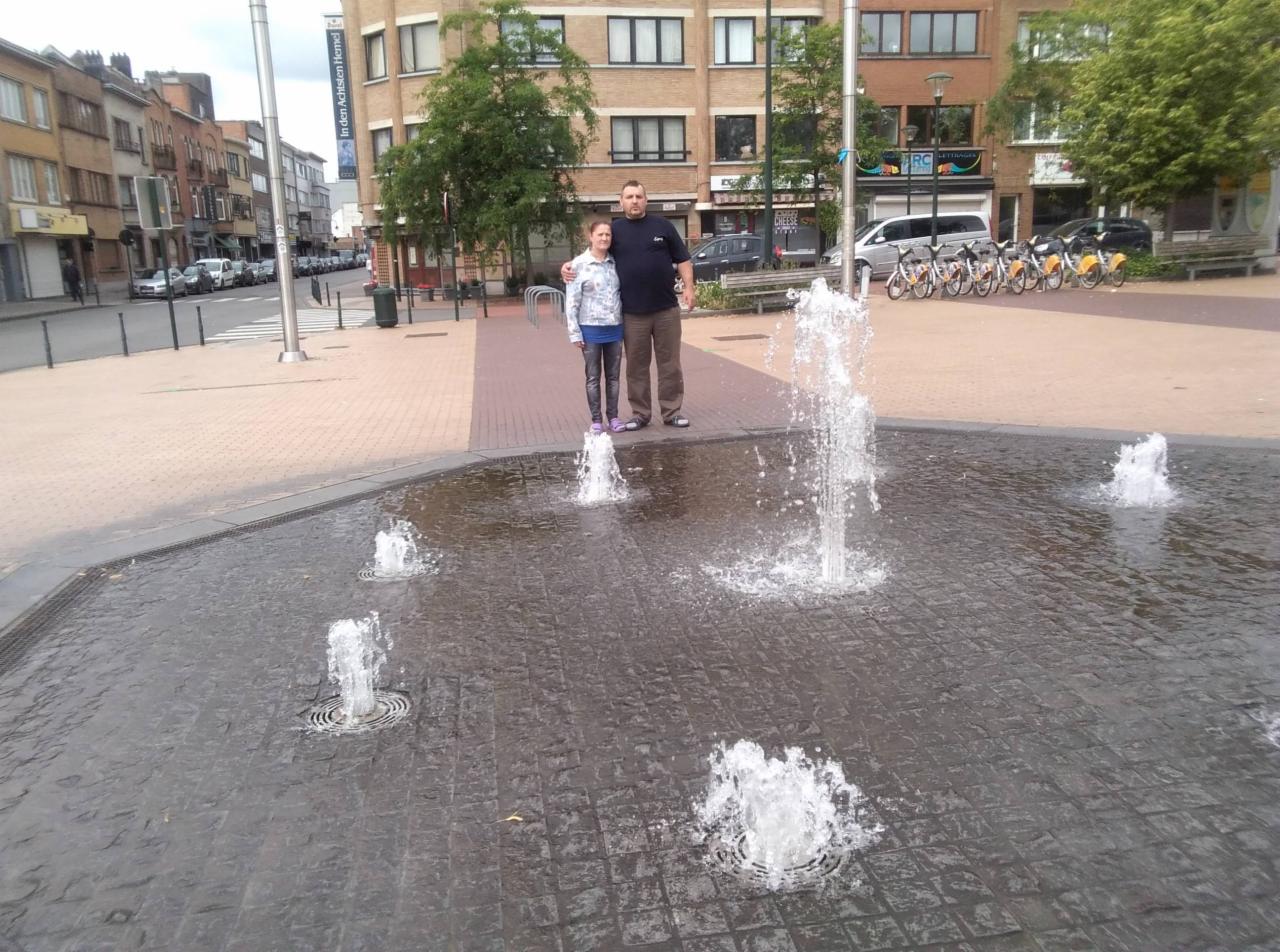 Para stoi na fontannie w mieście. W tle budynki, rowery miejskie i zaparkowane samochody. Ujęcie z poziomu fontanny, woda w ruchu. Lekkie zachmurzenie.