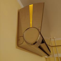 Klimatyzacjelegnica.pl - Nowoczesny klimatyzator ścienny w beżowym odcieniu, zamontowany w rogu pokoju, z widoczną kratką wentylacyjną i fragmentem suszarki na ubrania w tle.