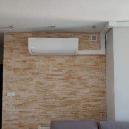 Klimatyzacjelegnica.pl - Biały klimatyzator Kaisaier zamontowany na ścianie z dekoracyjnej cegły w salonie, widoczne białe listwy maskujące instalację, w tle fragment kuchni.