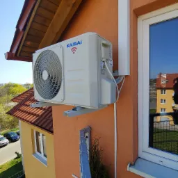 Zewnętrzna jednostka klimatyzacji Kaisai zamontowana na ścianie pomarańczowego budynku z widocznym oknem i fragmentem dachu, z przewodami w białej listwie.