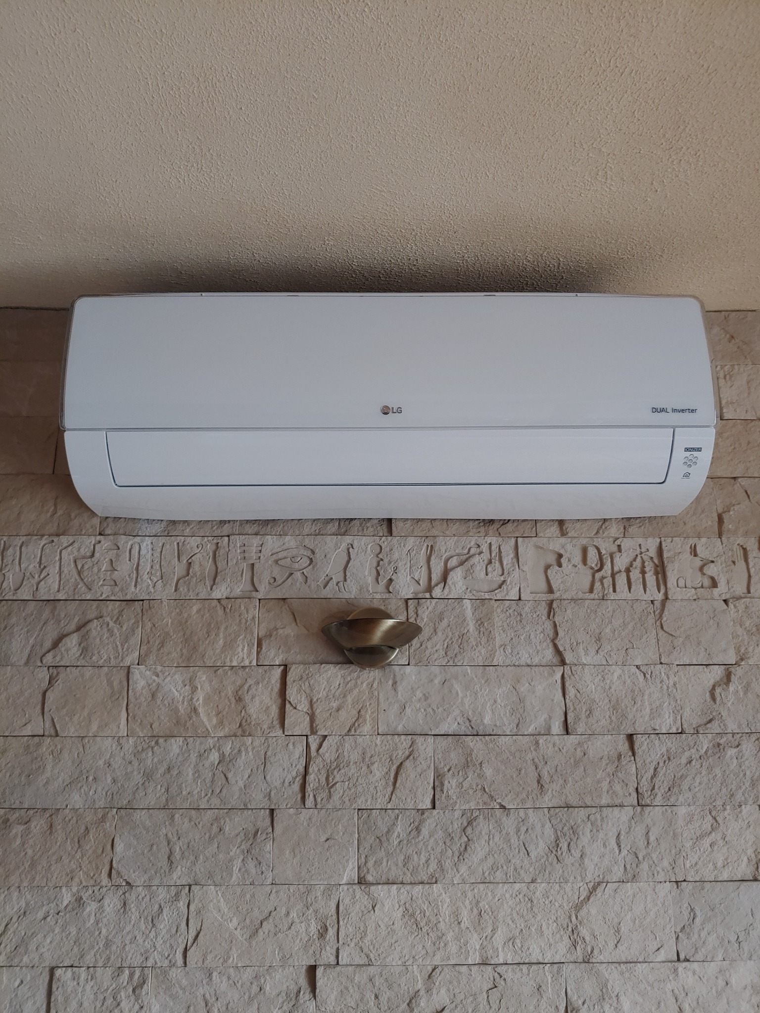 Biały klimatyzator LG Dual Inverter zamontowany na ścianie z dekoracyjnej cegły z motywem hieroglifów, podświetlony kinkietem.