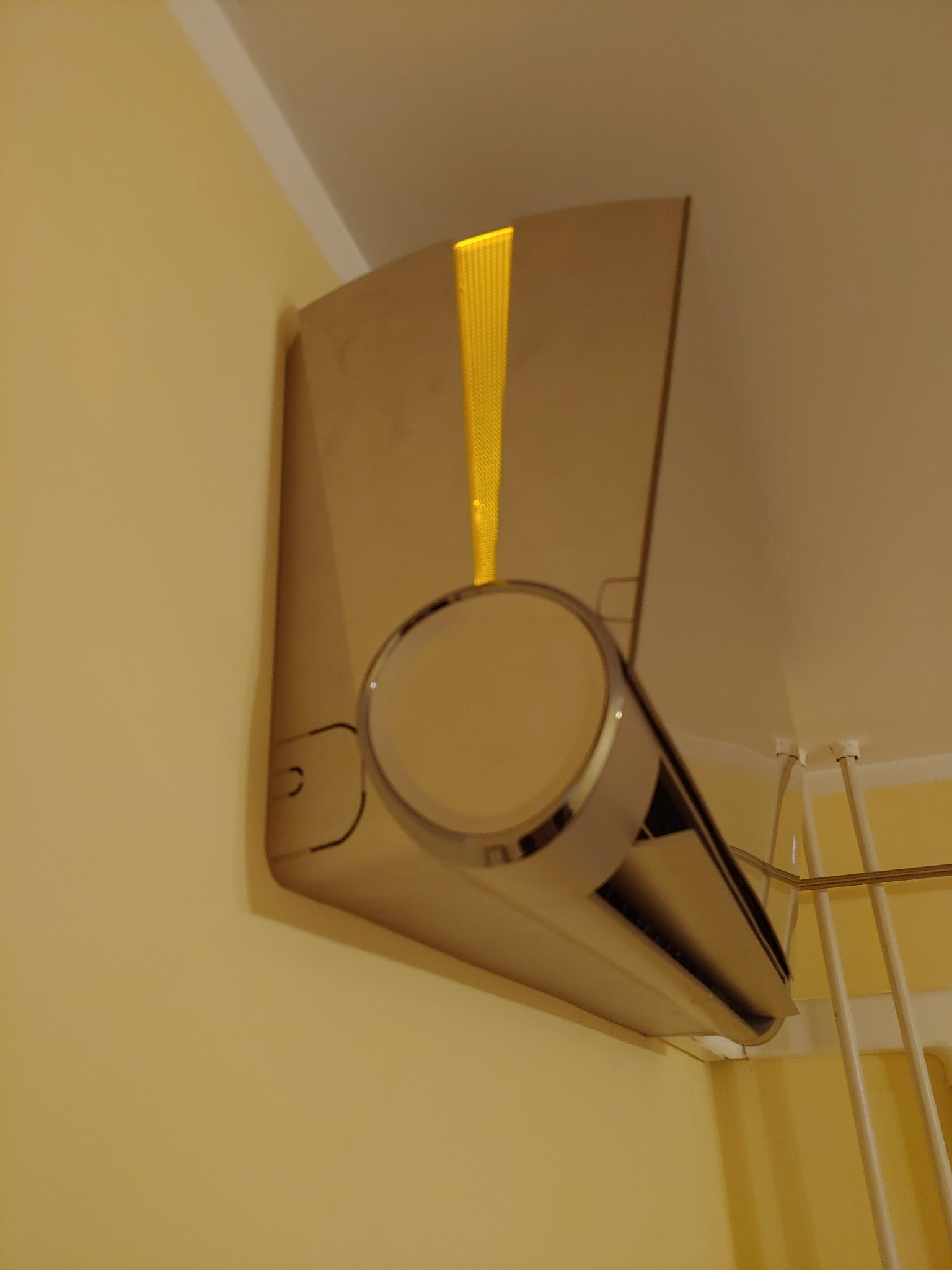 Nowoczesny klimatyzator ścienny w beżowym odcieniu, zamontowany w rogu pokoju, z widoczną kratką wentylacyjną i fragmentem suszarki na ubrania w tle.