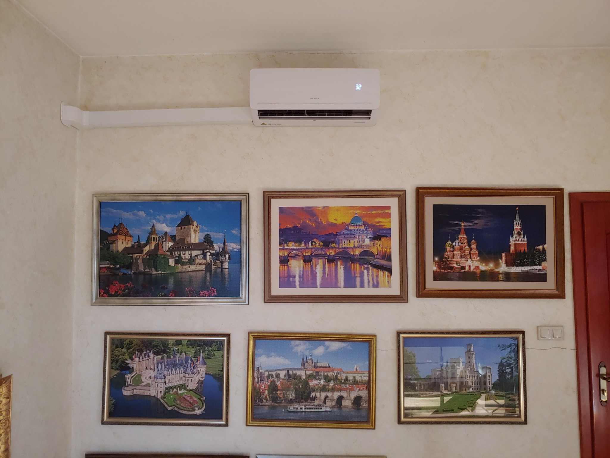 Biały klimatyzator ścienny marki Sevra, ustawiony nad rzędem sześciu obrazów z motywami architektonicznymi i krajobrazowymi w ozdobnych ramach, zamontowany na kremowej ścianie o fakturze...