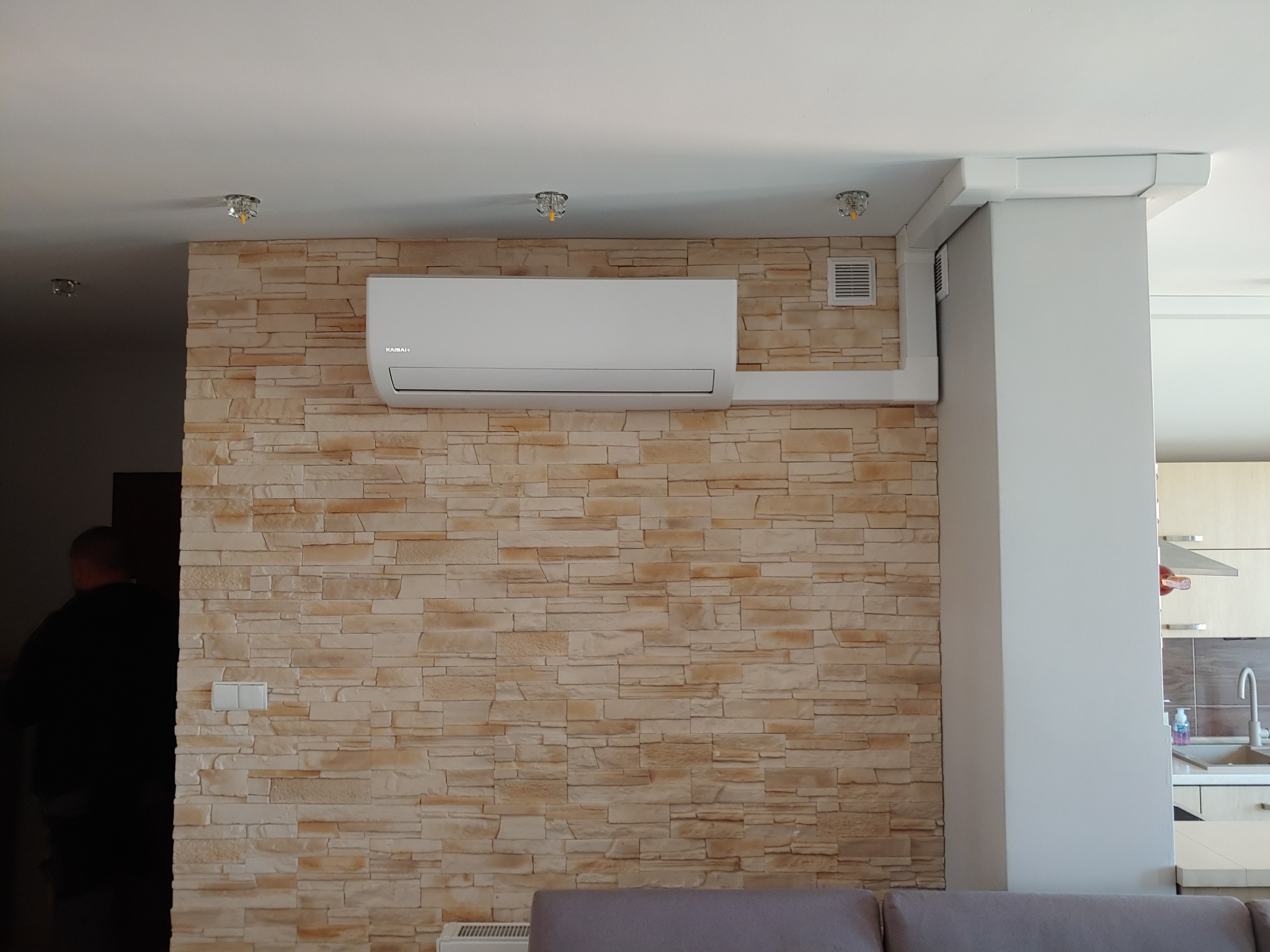 Biały klimatyzator Kaisaier zamontowany na ścianie z dekoracyjnej cegły w salonie, widoczne białe listwy maskujące instalację, w tle fragment kuchni.