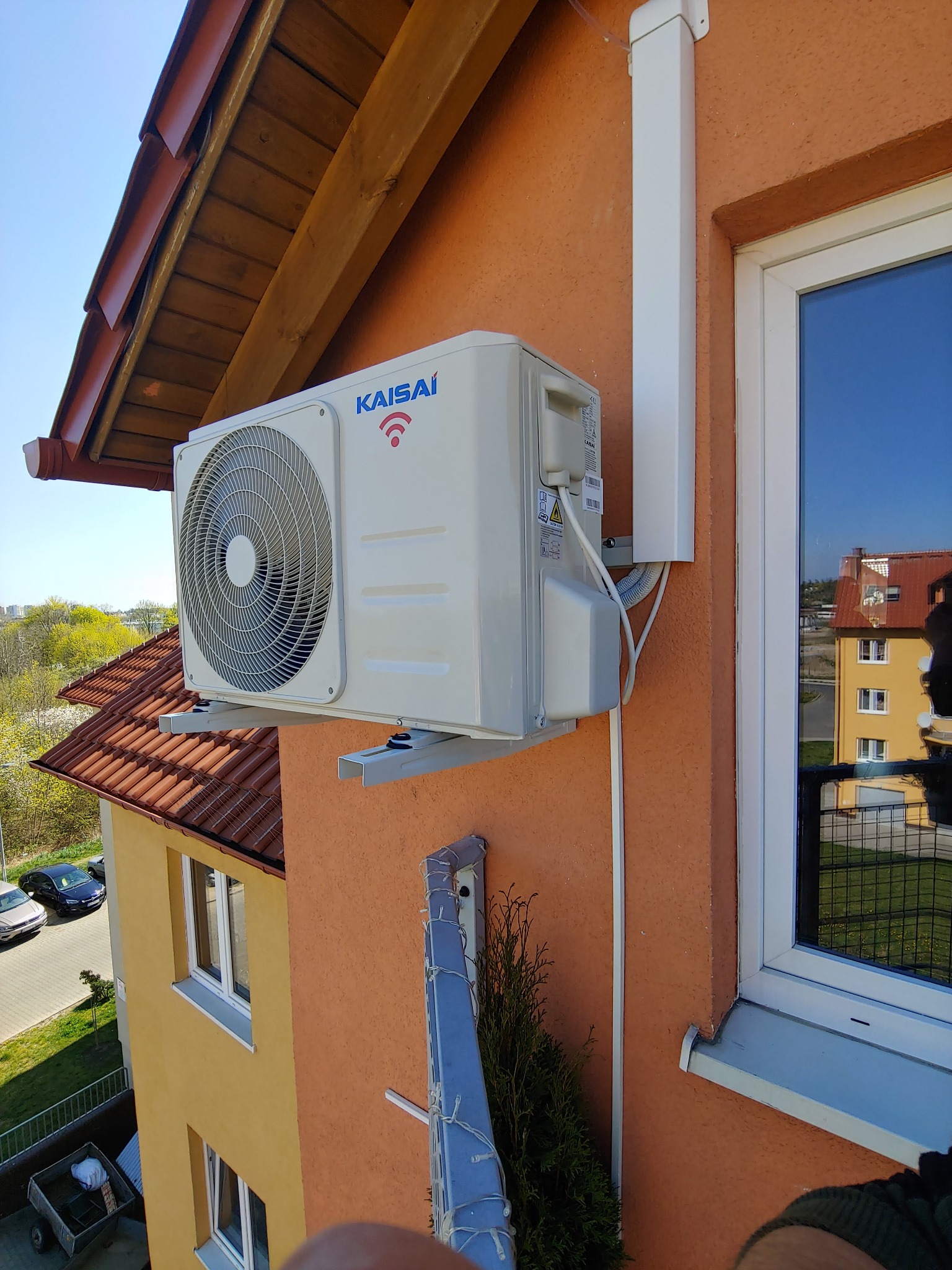 Zewnętrzna jednostka klimatyzacji Kaisai zamontowana na ścianie pomarańczowego budynku z widocznym oknem i fragmentem dachu, z przewodami w białej listwie.