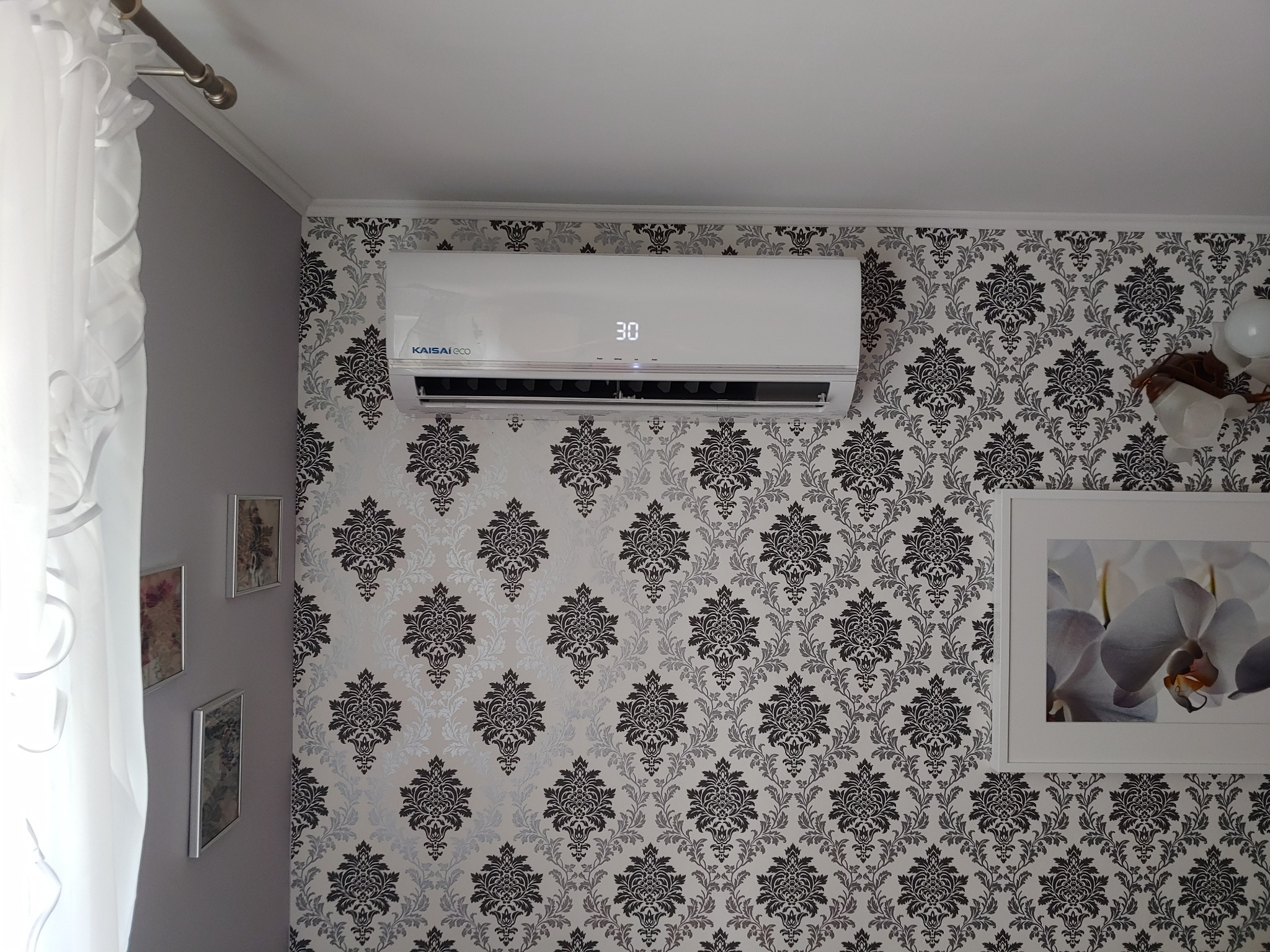 Biały klimatyzator marki Kaisai Eco zamontowany na ścianie z wzorzystą tapetą w salonie, wyświetlający temperaturę 30 stopni Celsjusza.
